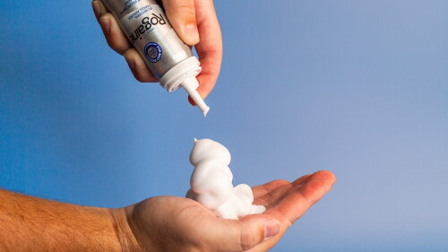 Uma mão segurando o tratamento com espuma de minoxidil Rogaine®, mostrando a aplicação do produto como parte de uma rotina diária de recrescimento de cabelo.